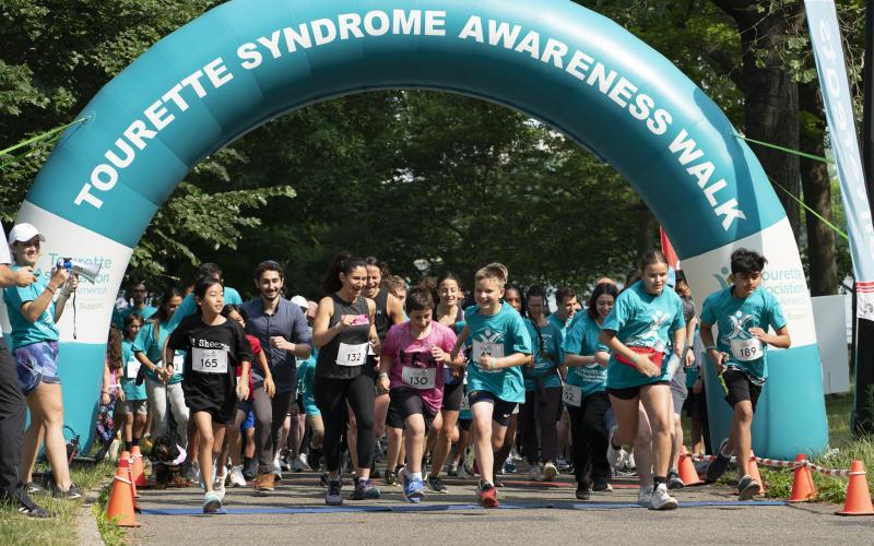 People running under a teal arch that says "Tourette Syndrome Awareness Walk"